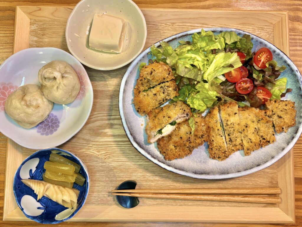 サッカー観戦後の晩ごはん全体。青紫蘇チーズ挟みミラノ風カツレツ、野菜サラダ、ふきと筍の煮物、冷奴、老祥記の豚まんが木製トレーに並ぶ