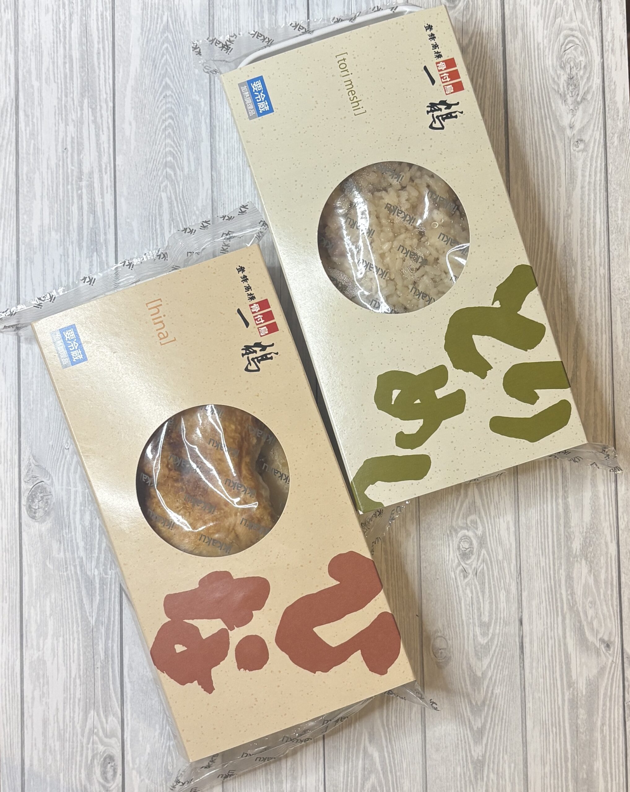 香川の骨付き鳥専門店・一鶴のお取り寄せセット。骨付き鳥ひな（手前）ととり飯（奥）の2点パッケージを並べて撮影。窓から中身が見えるデザインの化粧箱入り冷凍商品。