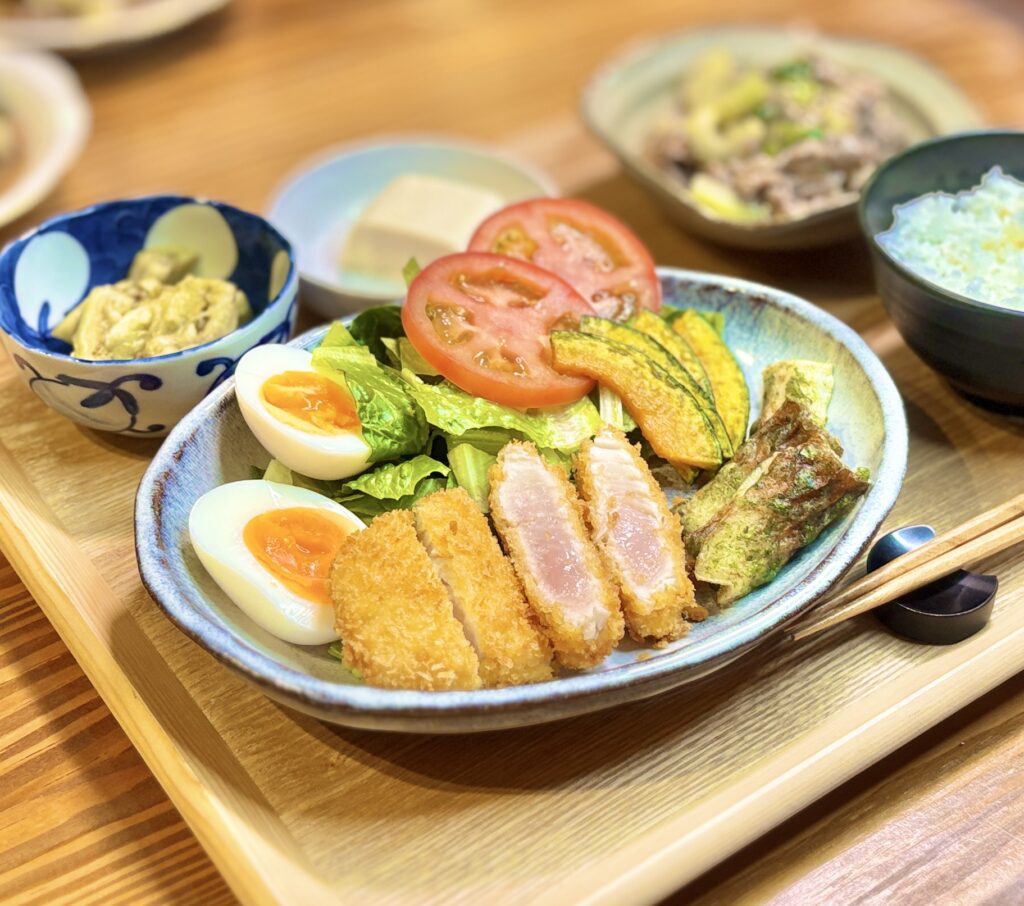 ビンチョウマグロのレアカツをメインにしたワンプレート定食の写真。塩麹パウダーで下味をつけたマグロカツは中がレアでしっとりとした仕上がり。ロメインレタスとトマト、半熟ゆで卵、かぼちゃの素揚げを添えたサラダ、竹輪の磯辺揚げも盛り付けられており、バランスの良い献立になっている。奥にはセロリと牛肉の中華炒め、焼きなす、冷奴、ごはんも並ぶ家庭料理の食卓。