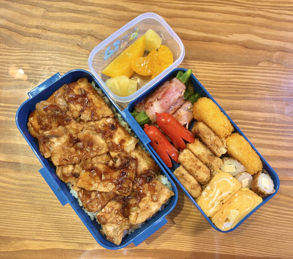 木目のテーブルの上に置かれた二段弁当箱の写真。下段には白ごはんの上に甘辛ダレと刻みネギをかけた鶏肉がたっぷりのせられ、上段にはウインナー、ベーコン巻きアスパラ、コロッケ、チキンナゲット、卵焼きなどのおかずが彩りよく詰められている。別容器にはみかん、パイナップル、黄桃などのフルーツが入ったバランスの良い手作り弁当。