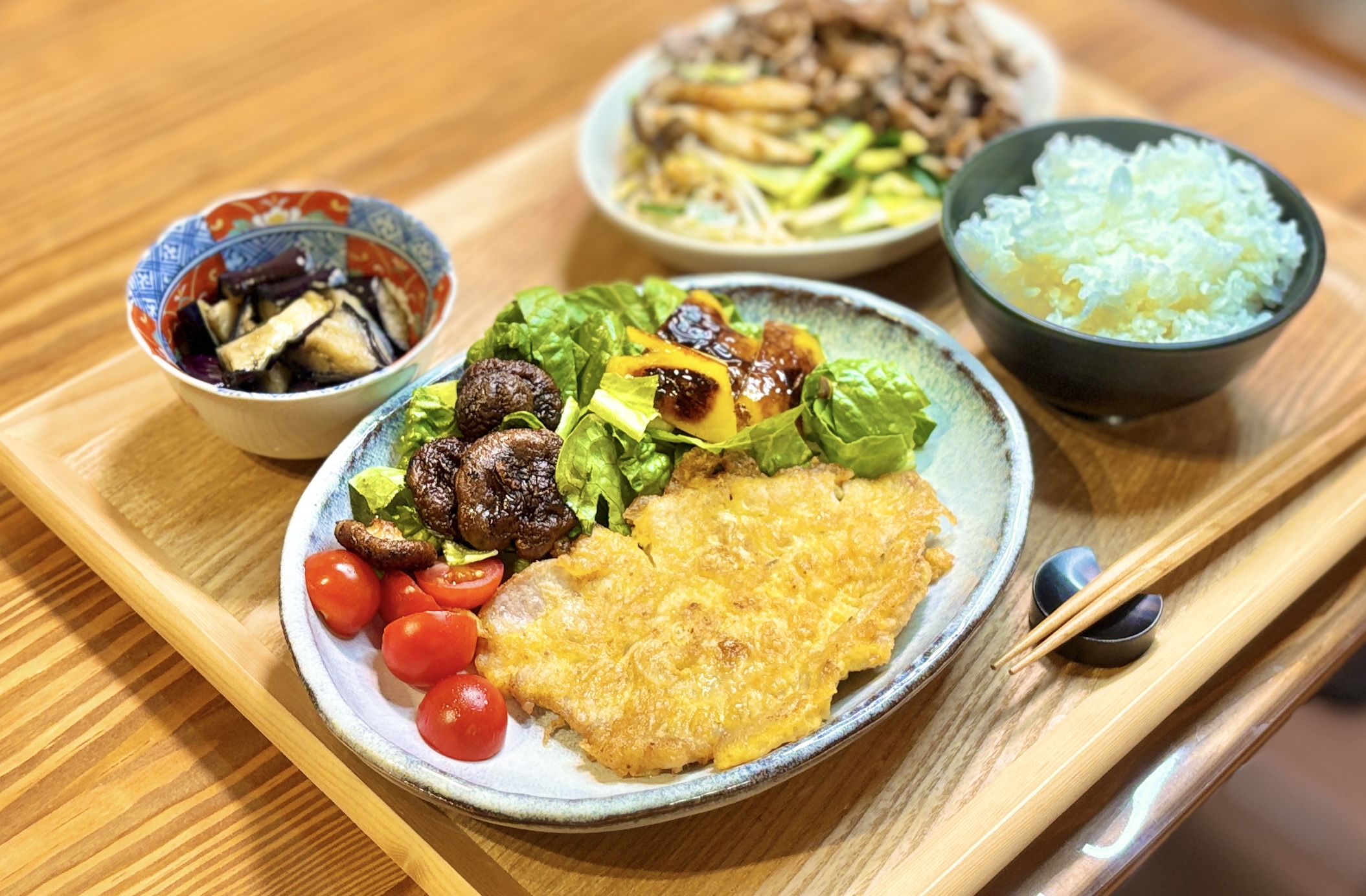 旦那キッチンの晩ごはん全体。手前にポークピカタと焼き椎茸・焼きパプリカ・ロメインレタス・ミニトマトのサラダを盛った大皿、左奥に茄子の揚げ浸しの小鉢、右奥にジンギスカンのお皿とご飯茶碗が並ぶ、ビールに合う家庭料理の献立