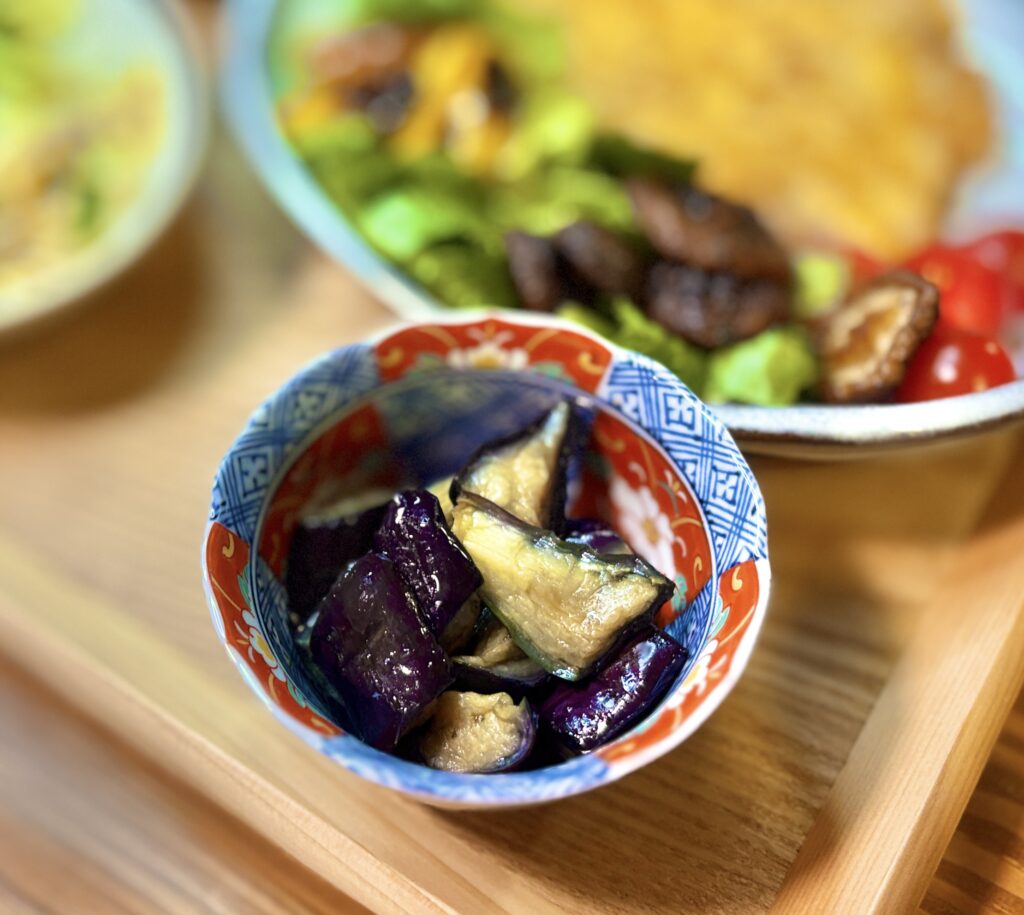 旦那キッチンの定番小鉢、茄子の揚げ浸しのクローズアップ。艶やかな紫色の茄子がだし汁にじんわり浸かり、伊万里風の赤絵の小鉢に盛り付けられている。奥にはポークピカタのサラダ皿がぼんやりと見える、ビールに合う和の副菜レシピ