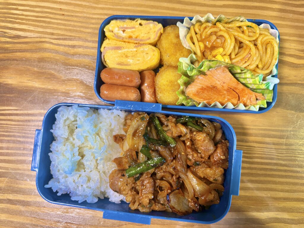 木目のテーブルの上に置かれた二段弁当箱の写真。下段には白ごはんと、玉ねぎとニラ入りの甘辛い豚肉炒めがたっぷり詰められている。上段には卵焼き、ウインナー、コロッケ、焼き鮭、ナポリタンが彩りよく盛り付けられた、ボリューム満点の手作り弁当。