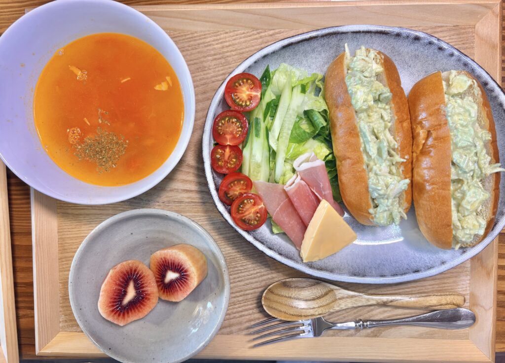木製トレーに盛られた旦那キッチンの休日ランチプレート。チキンアボカドクリームチーズワサビマヨ和えのコッペパンサンド、生ハム、レッドチェダーチーズ、プチトマト、きゅうり入りのサラダ、トマトミネストローネ、レッドキウイが並ぶカフェ風の一皿。