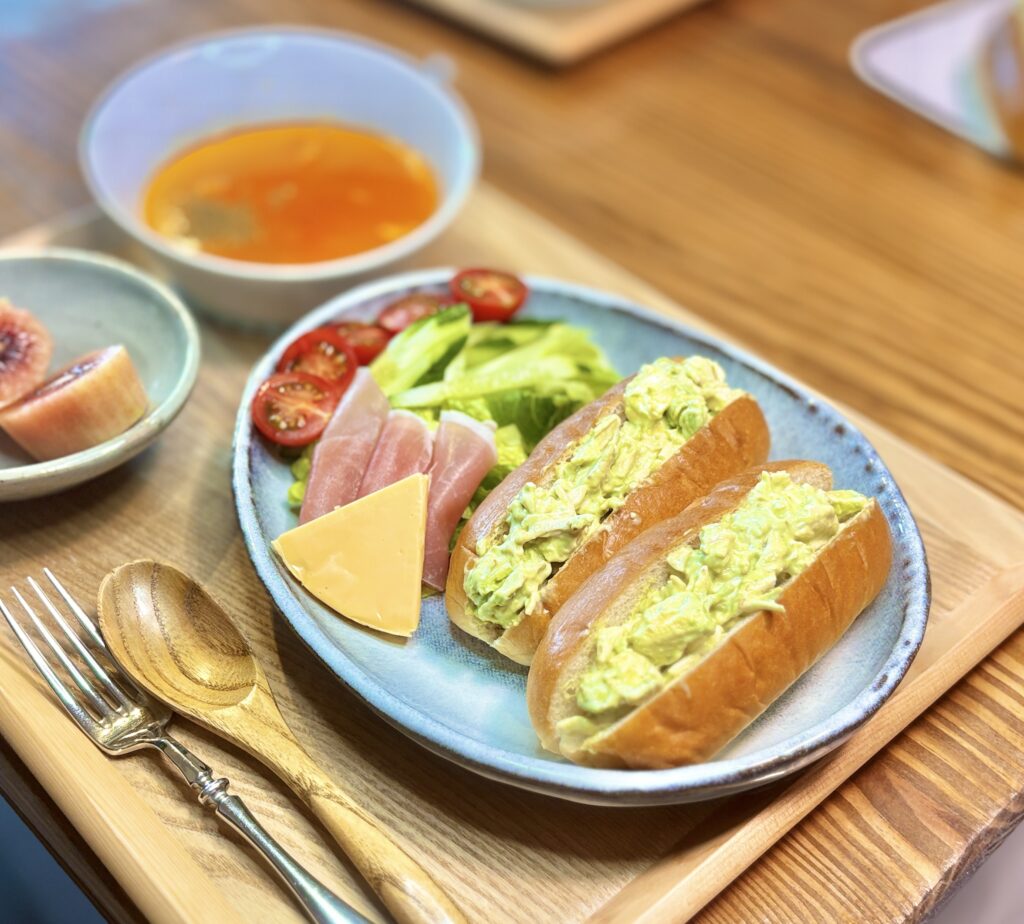 コッペパンにたっぷりのせたチキンアボカドクリームチーズワサビマヨ和えのサンド。淡い黄緑色の具材がなみなみと盛られ、隣には生ハム、チェダーチーズ、プチトマト、きゅうり、レタスが彩りよく並ぶ。