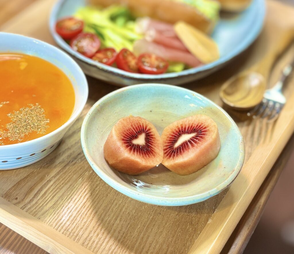 淡いターコイズブルーの小皿に置かれたレッドキウイの断面。濃い赤〜えんじ色のグラデーションが美しく、白いラインが中央を走っている。背景にはミネストローネとサラダプレートがぼんやりと見える。