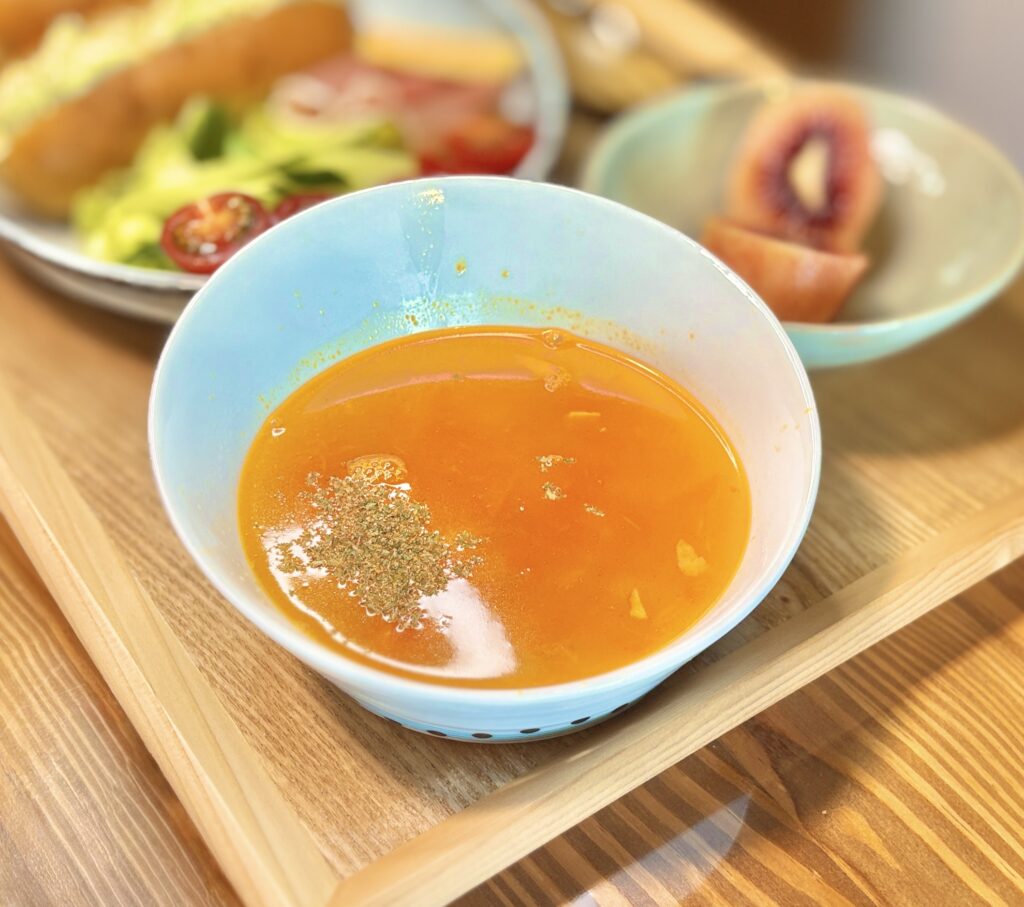 ターコイズブルーのスープボウルに入ったトマトミネストローネ。オレンジ色のトマトベースのスープに、パプリカ、ズッキーニ、大根、ベーコン、茄子の具材がたっぷり入り、乾燥ハーブが散らされている。