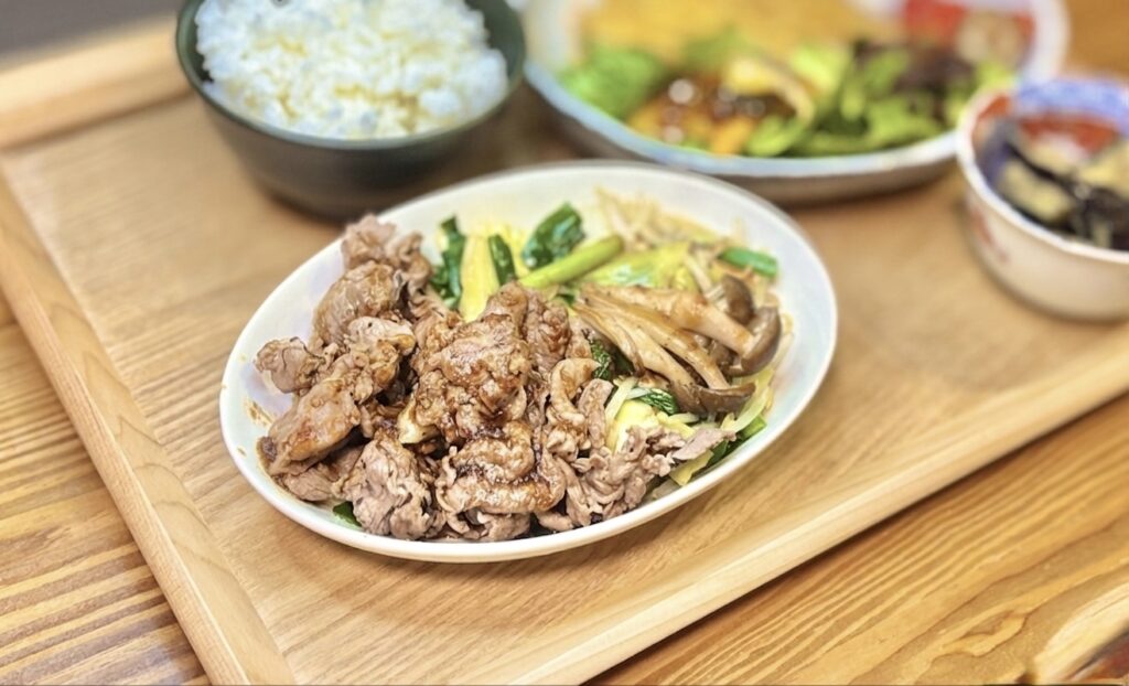 旦那キッチンのジンギスカンのクローズアップ。タレがしっかり絡んだジューシーなラム薄切り肉が白い楕円皿に盛られ、もやし・キャベツ・ニラ・舞茸と一緒に炒められている。奥にはご飯茶碗とポークピカタのサラダ皿、茄子の揚げ浸しの小鉢が見える、ビールに合うラム肉レシピの晩ごはん