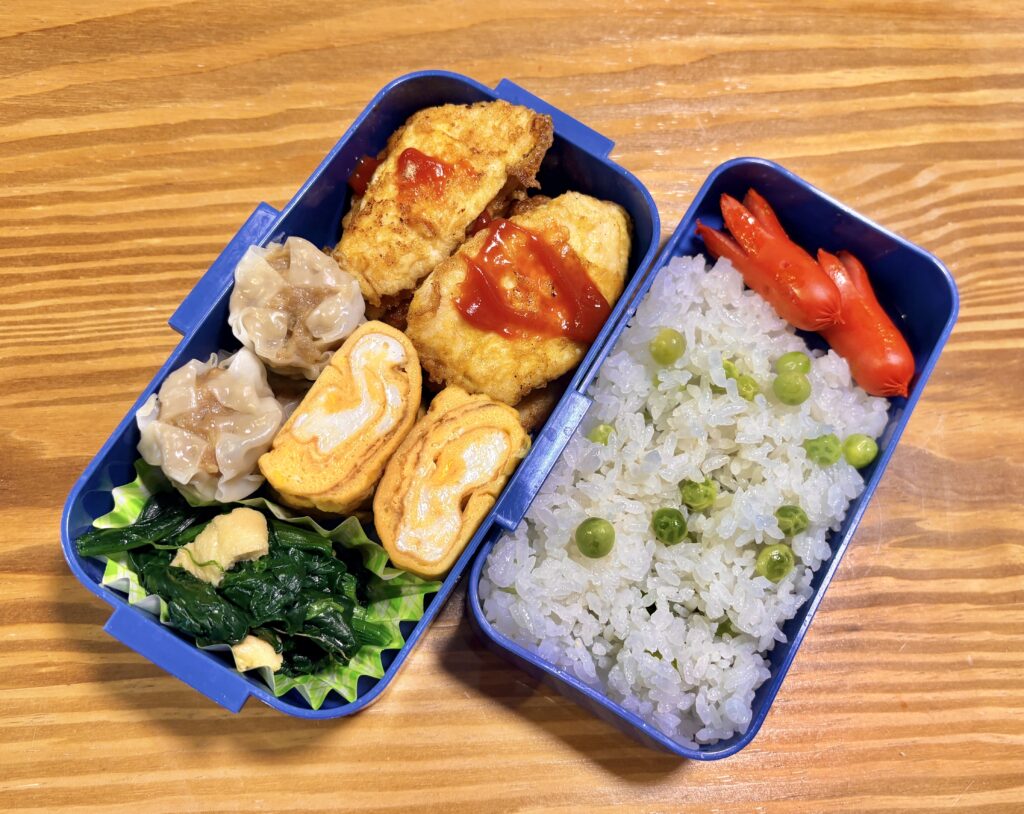 木目のテーブルの上に置かれた二段弁当箱の写真。右側にはグリーンピースごはんとタコさんウインナー、左側にはケチャップをかけたチキンソテー、焼売、卵焼き、ほうれん草のおひたしがバランスよく詰められている、彩り豊かな手作り弁当。