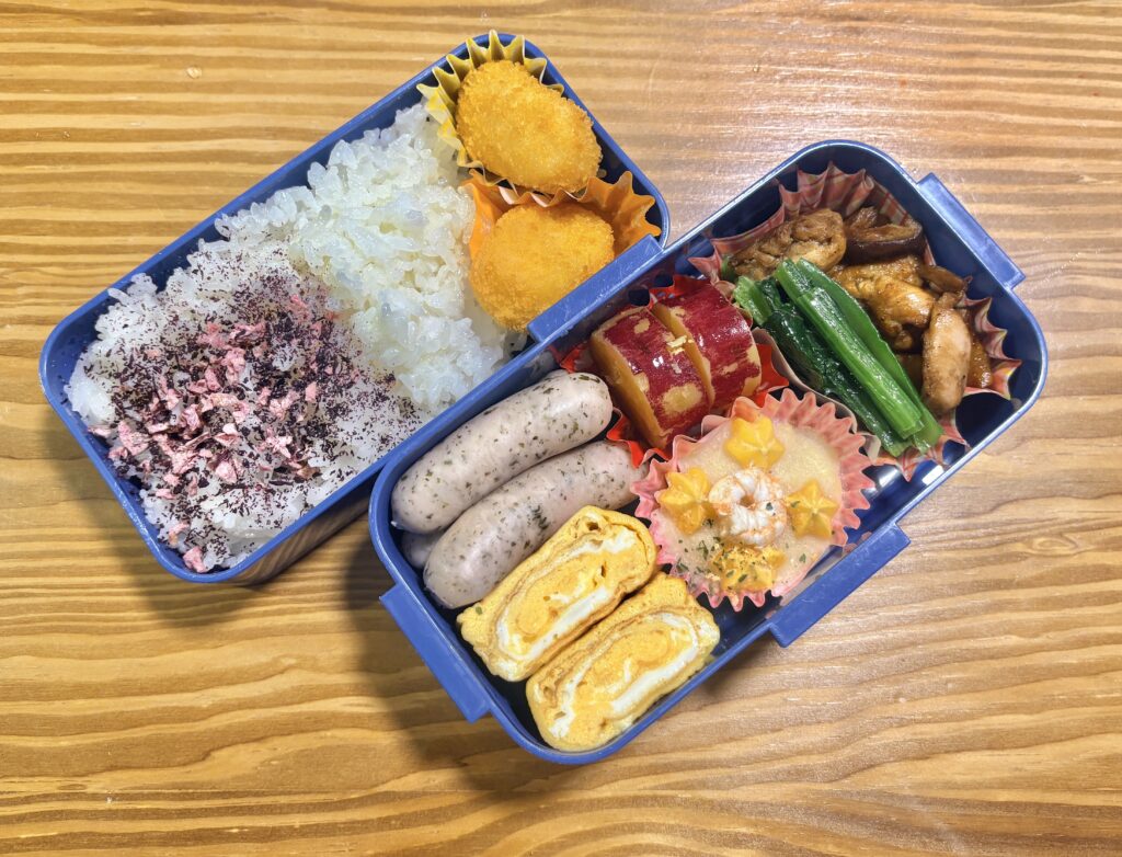 木目のテーブルの上に置かれた二段弁当箱の写真。左側には白ごはんにふりかけをかけ、コロッケが添えられている。右側には卵焼き、ソーセージ、大学いも、鶏肉と野菜の炒め物、ほうれん草のおひたし、エビ入りのおかずが彩りよく詰められた、栄養バランスの良い手作り弁当。