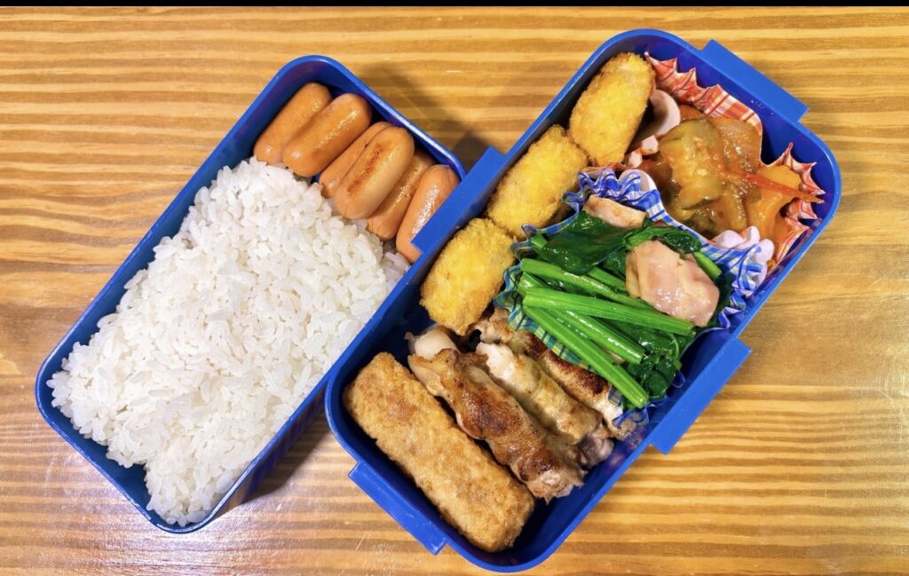木目のテーブルの上に置かれた二段弁当箱の写真。左側には白ごはんとウインナーが並び、右側にはコロッケ、ちくわの磯辺揚げ、焼き鳥、ほうれん草のおひたし、野菜の煮物などのおかずが彩りよく詰められている、ボリューム満点の手作り弁当。