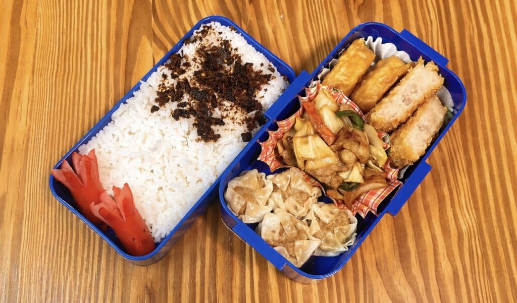 木目のテーブルの上に置かれた二段弁当箱の写真。左側には白ごはんにふりかけとタコさんウインナーが添えられ、右側には焼売、野菜と豚肉の炒め物、コロッケや揚げ物が彩りよく詰められている、ボリューム満点の手作り弁当。