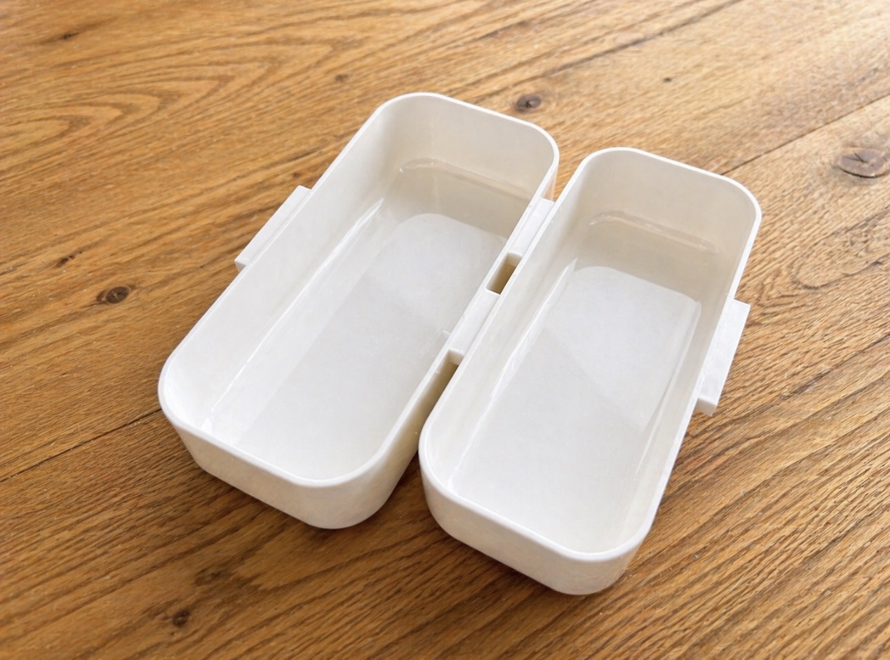 木目が美しいナチュラルな木製テーブルの上に、空の白い二段弁当箱が開いた状態で置かれている様子を斜め前から撮影した写真。シンプルで清潔感のある白い弁当箱は仕切り付きで、これからお弁当を詰める前の準備シーンやお弁当作りのイメージに最適な構図。