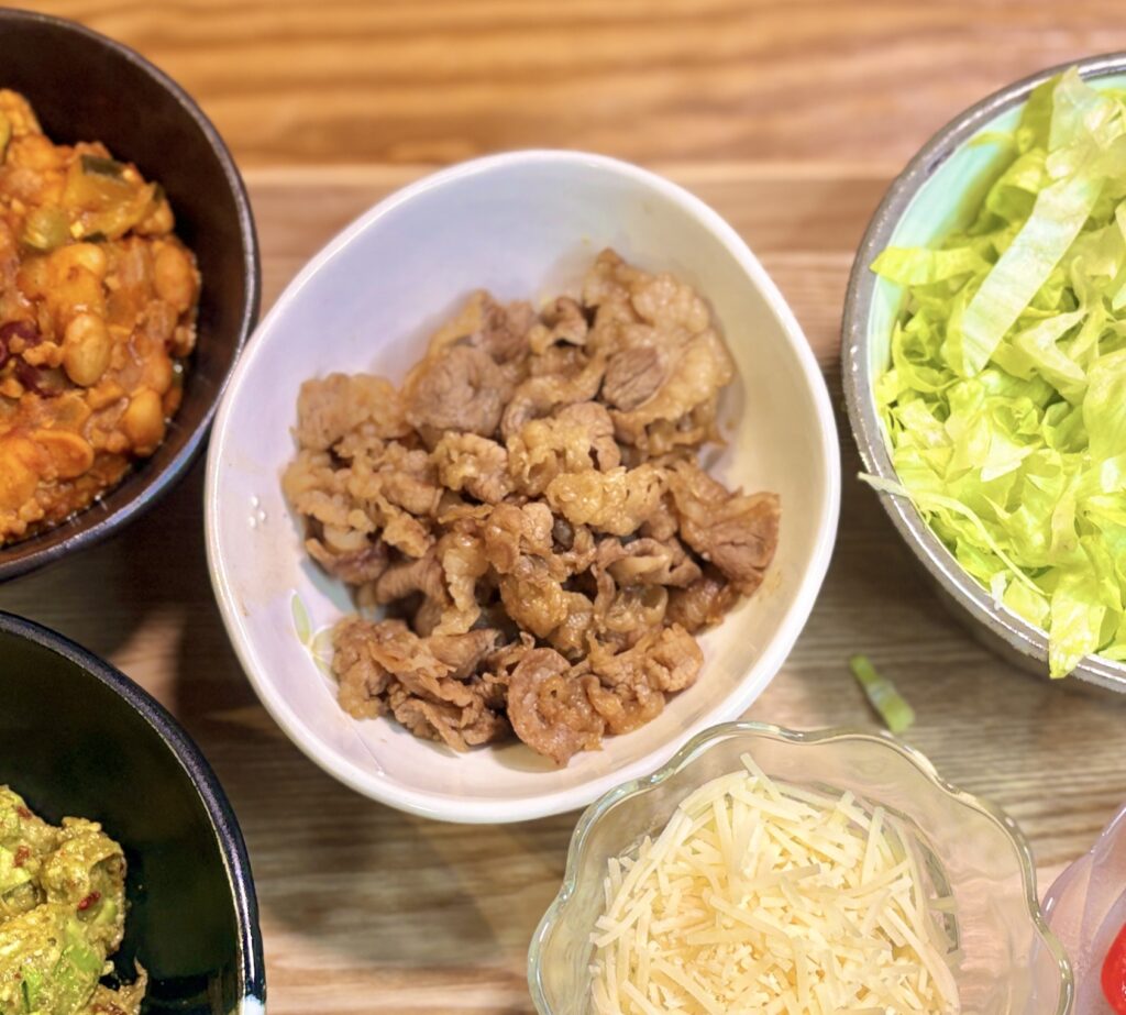 木製テーブルに並んだタコスの具材一式。中央の白いボウルに牛肉のすき焼き風、左上の黒いボウルにひき肉と豆のトマトチリ煮込み、右上にざく切りレタス、左下にワカモレ、下中央に十勝チーズの細切りが盛られている。