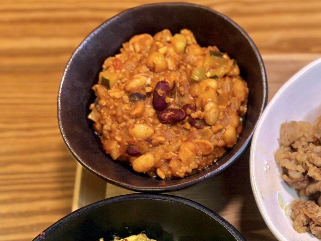 茶色い丸いボウルに盛られたひき肉と豆のトマトチリ煮込み。大豆、金時豆、ひき肉がトマトベースのソースと絡まり、ズッキーニも混ざった具だくさんの一品。