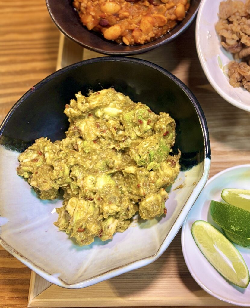 黒と白のグラデーションが美しい陶器のボウルに盛られたワカモレ。アボカドを粗くつぶしてワカモレシーズニングで和えた本格グアカモレで、右下にはライムのくし切りが添えられている。奥にはひき肉と豆のトマトチリ煮込みと牛肉のすき焼き風も見える。