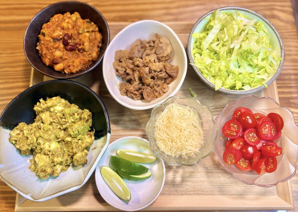 木製トレーに並んだタコスの具材一式。ひき肉と豆のトマトチリ煮込み、牛肉のすき焼き風、ワカモレ、千切りレタス、十勝チーズ、ライム、ミニトマトがそれぞれ小鉢に盛られたおうちタコスパーティーの準備風景。