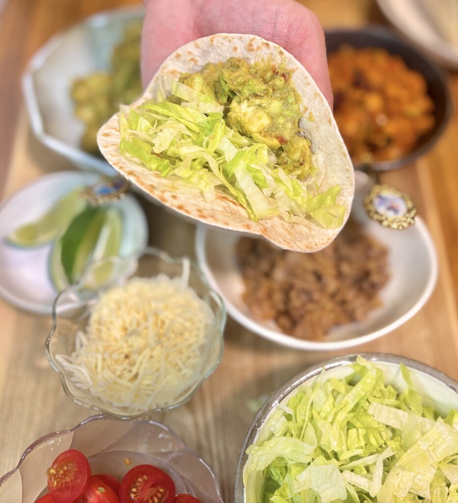 手でトルティーヤを持ち、ワカモレとレタスをのせて巻いているところ。背後にはひき肉チリ豆煮込みや十勝チーズ、ミニトマトなどの具材が並ぶ。