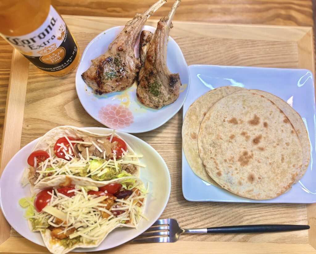 木製トレーに並んだ旦那キッチンのメキシカンナイト全景。左下に十勝チーズとミニトマトをたっぷりのせたタコス、中央上に骨付きラムチョップのグリル、右にフライパンで焼いたトルティーヤ、左上にコロナビールが並ぶ。