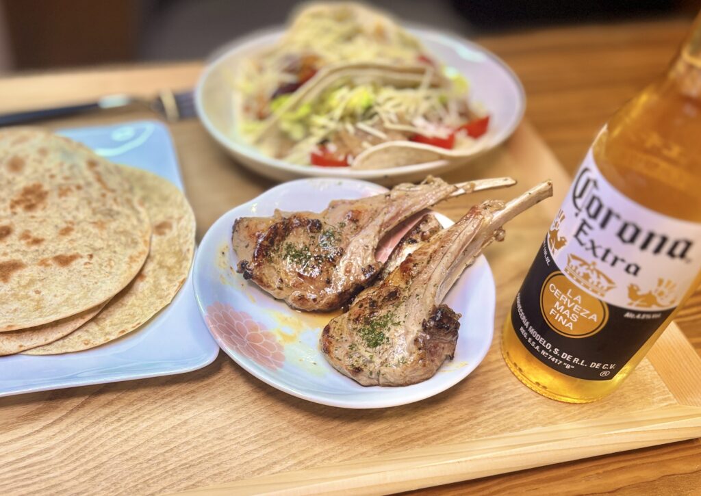 花柄の白い小皿に盛られた骨付きラムチョップのグリル。こんがりと焼き色がついてパセリが散らされており、隣にはフライパンで焼いたトルティーヤとコロナビールが並ぶ。