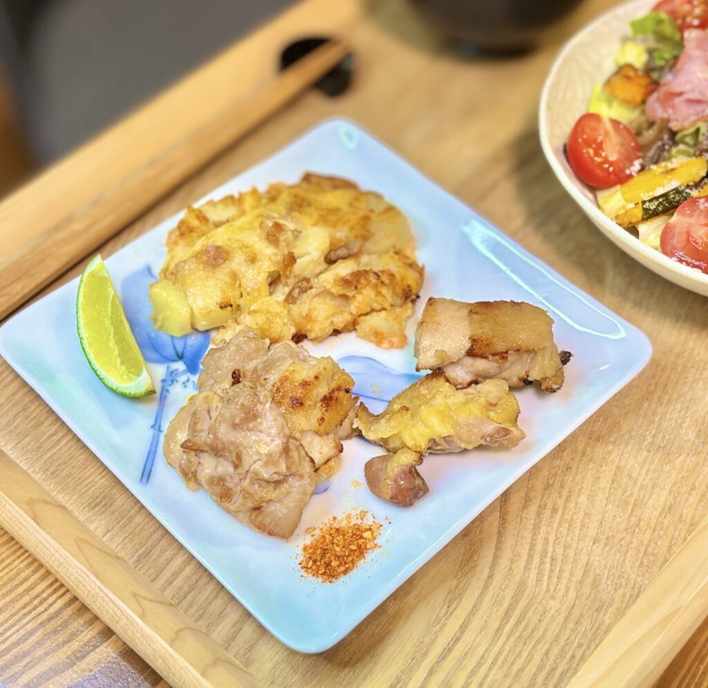 名古屋コーチンと国産若鶏を粉塩麹で焼いた食べ比べプレート。香ばしく焼き上げた塩麹チキンにライムを添え、同じ皿にはツナとチーズとポテトを混ぜてフライパンで焼いたポテト焼きも盛り付け。木製トレーの上に並んだ、白ワインに合う鶏肉料理と副菜の晩ごはん。