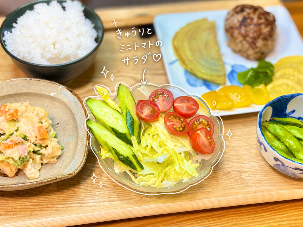 ガラスの器に、きゅうり、ミニトマト、レタスを盛り付けたシンプルな野菜サラダ。白い手書き風の文字入れで、きゅうりとミニトマトのサラダと可愛く紹介しています。赤いミニトマトと緑のきゅうりが鮮やかで、ハンバーグや焼き野菜と一緒に並べると食卓が明るくなる副菜です。ハンバーグ献立に合うさっぱりサラダ、簡単な野菜副菜、彩りのよいおうちごはんの盛り付け例として使える写真です。