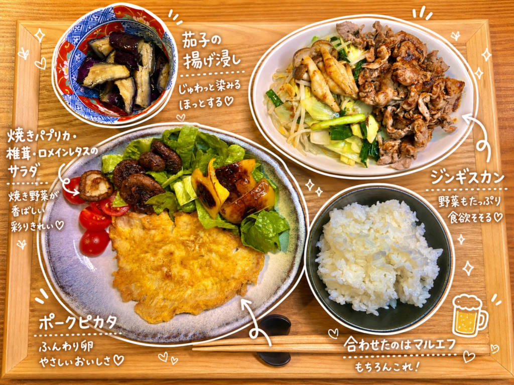 旦那キッチンのジンギスカンとポークピカタの晩ごはん全体を真上から撮影。手前左にポークピカタと焼き椎茸・焼きパプリカ・ロメインレタス・ミニトマトのサラダ皿、左奥に茄子の揚げ浸しの伊万里風小鉢、右奥にジンギスカンのラム肉炒め皿とご飯茶碗が木製トレーに並ぶ、ビールに合う家庭料理の献立