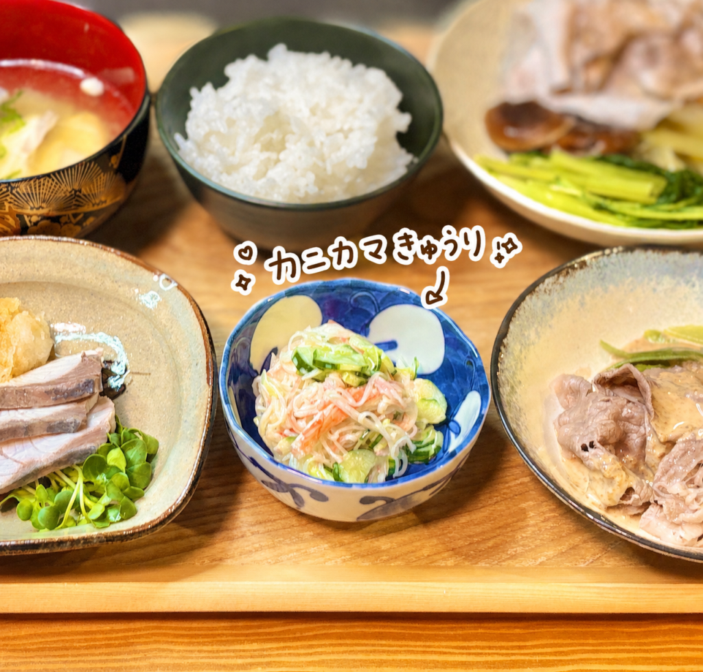 カニカマときゅうりをマヨネーズで和えた、簡単副菜の写真です。中央の青い小鉢には、ほぐしたカニカマと薄切りきゅうりが彩りよく盛りつけられており、マヨネーズで和えたまろやかな味わいと、きゅうりのシャキッとした食感が伝わります。写真には、カニカマきゅうりという可愛い手書き風の文字入れと矢印が入り、小鉢の副菜をわかりやすく紹介しています。周りには、ぶりとカイワレのおろししゃぶしゃぶ、黒毛和牛のゴマだれ、豚ロースのポン酢、白ごはんなどが写っていて、しゃぶしゃぶ献立の箸休めとしてぴったりです。カニカマきゅうり、きゅうり副菜、マヨネーズ和え、簡単小鉢、和食献立、おうちごはんブログに適した代替テキストです。
