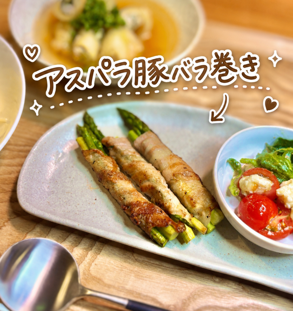 アスパラ豚バラ巻きを主役にした、旦那キッチン晩ごはんの一品写真です。淡いブルーの器には、こんがり焼き目のついたアスパラ豚バラ巻きが並び、豚バラ肉の香ばしさとアスパラの鮮やかな緑が食欲をそそります。右側には、アイスプラントとトマト、花畑牧場のカッテージチーズを使ったカプレーゼ風の副菜も添えられており、肉料理のこってり感に爽やかな彩りを加えています。写真上部には、アスパラ豚バラ巻きという手書き風の可愛い文字入れと矢印、ハートやきらきらの装飾があり、料理ブログや家庭料理の献立紹介にぴったりの明るい雰囲気です。アスパラの豚バラ巻きレシピ、簡単おかず、豚肉巻き、晩ごはん献立、旦那キッチンの副菜として紹介する画像に適しています。