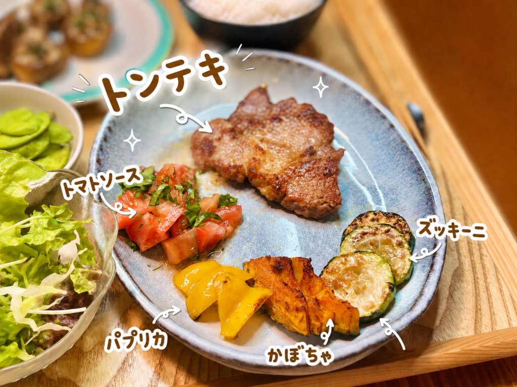 香ばしく焼いたトンテキに、刻んだトマトと紫蘇を合わせたさっぱりソース、ズッキーニ・かぼちゃ・パプリカの焼き野菜を添えた一皿。手書き風の文字入れでトンテキ用トマトソースと焼き野菜を紹介した、暑い日に食べやすいトンテキ献立の写真。