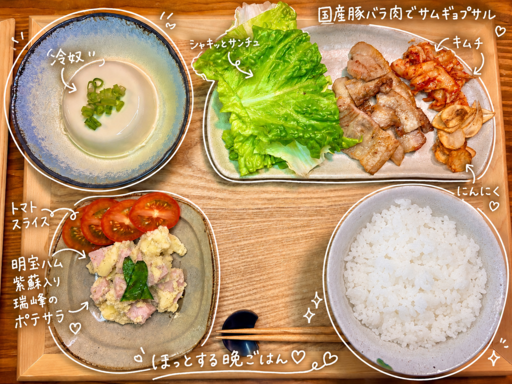 国産豚バラ肉で作ったサムギョプサルをメインに、シャキッとしたサンチュ、キムチ、焼きにんにく、冷奴、明宝ハム紫蘇入り瑞峰を使ったポテサラ、トマトスライス、白ごはんを木製トレーに並べた晩ごはん献立の写真。手書き風の文字入れで、サムギョプサルの具材や副菜がわかりやすく紹介されており、おうちで楽しむ韓国風ごはんと和の副菜を組み合わせた旦那キッチンの家庭料理メニュー。
