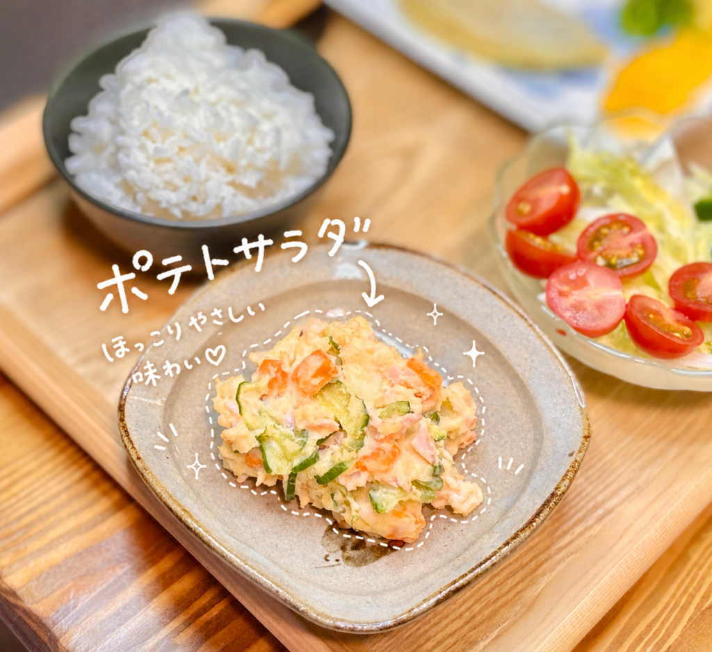 きゅうり、にんじん、ハムを混ぜ込んだ、ほっこりやさしい味わいのポテトサラダを小皿に盛り付けた副菜写真。手書き風の白文字でポテトサラダと紹介し、点線や星の飾りで可愛く文字入れしています。奥には白ごはんやミニトマトのサラダが見え、ハンバーグ献立の副菜としても相性のよい一品です。肉料理に合う副菜、家庭料理の定番ポテトサラダ、旦那キッチンのおうちごはん記録にぴったりの画像です。