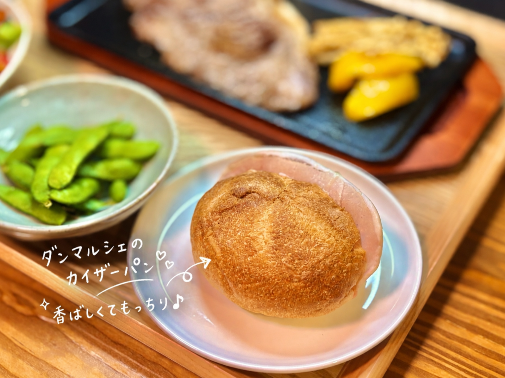ダンマルシェのカイザーパンを白い皿に盛り付けた、おうちディナーのパン写真。香ばしく焼き色のついた丸いカイザーパンにハムを合わせ、奥には岐阜県産枝豆やロースステーキの鉄板プレートが見える食卓風景。ステーキや具沢山シーザーサラダ、赤ワインと一緒に楽しむパンとして、洋食屋さんのような雰囲気を添えてくれる旦那キッチンのおうちごはんメニュー。
