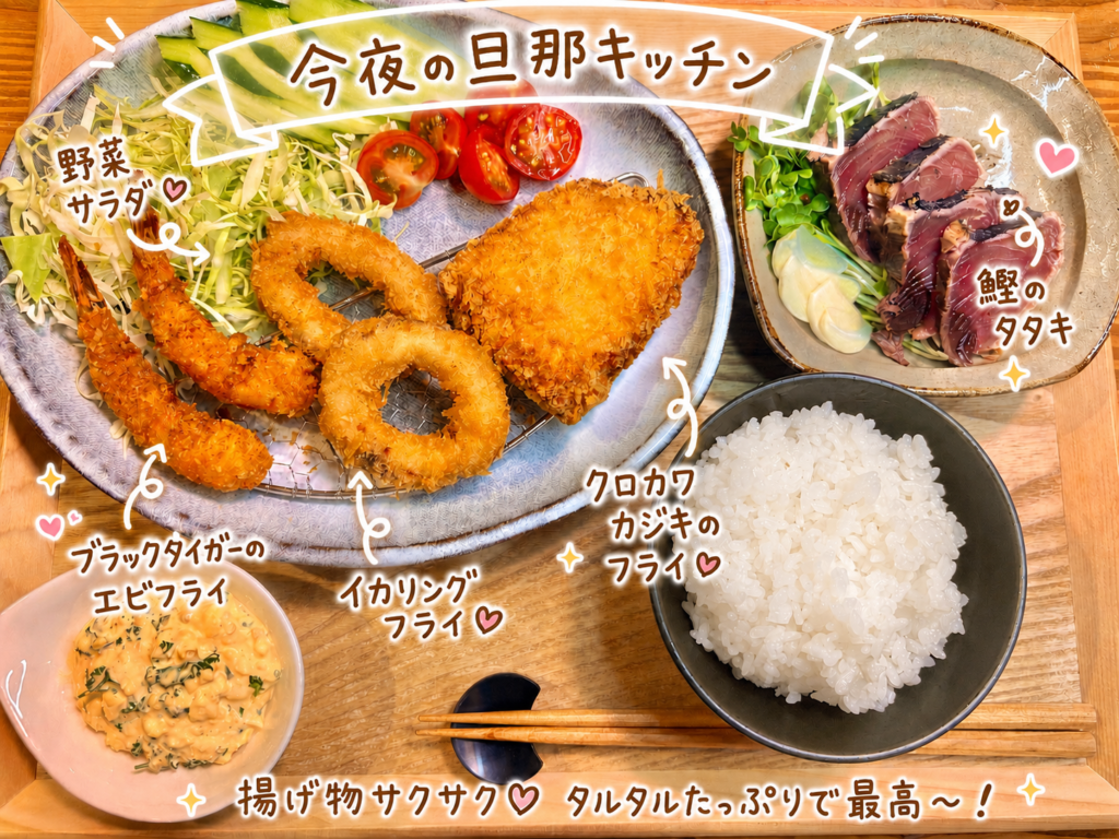 旦那キッチンの揚げ物だらけの晩ごはん全体俯瞰。手前の大皿にブラックタイガーのエビフライ・イカリングフライ・クロカワカジキのフライがサクサクの衣で勢ぞろい、キャベツの千切りときゅうり・ミニトマトの野菜サラダを添えて。手前には手作りタルタルソースの小鉢、右奥に鰹のタタキとご飯茶碗が並ぶ、白ワインに合うフライ三種盛り献立。
