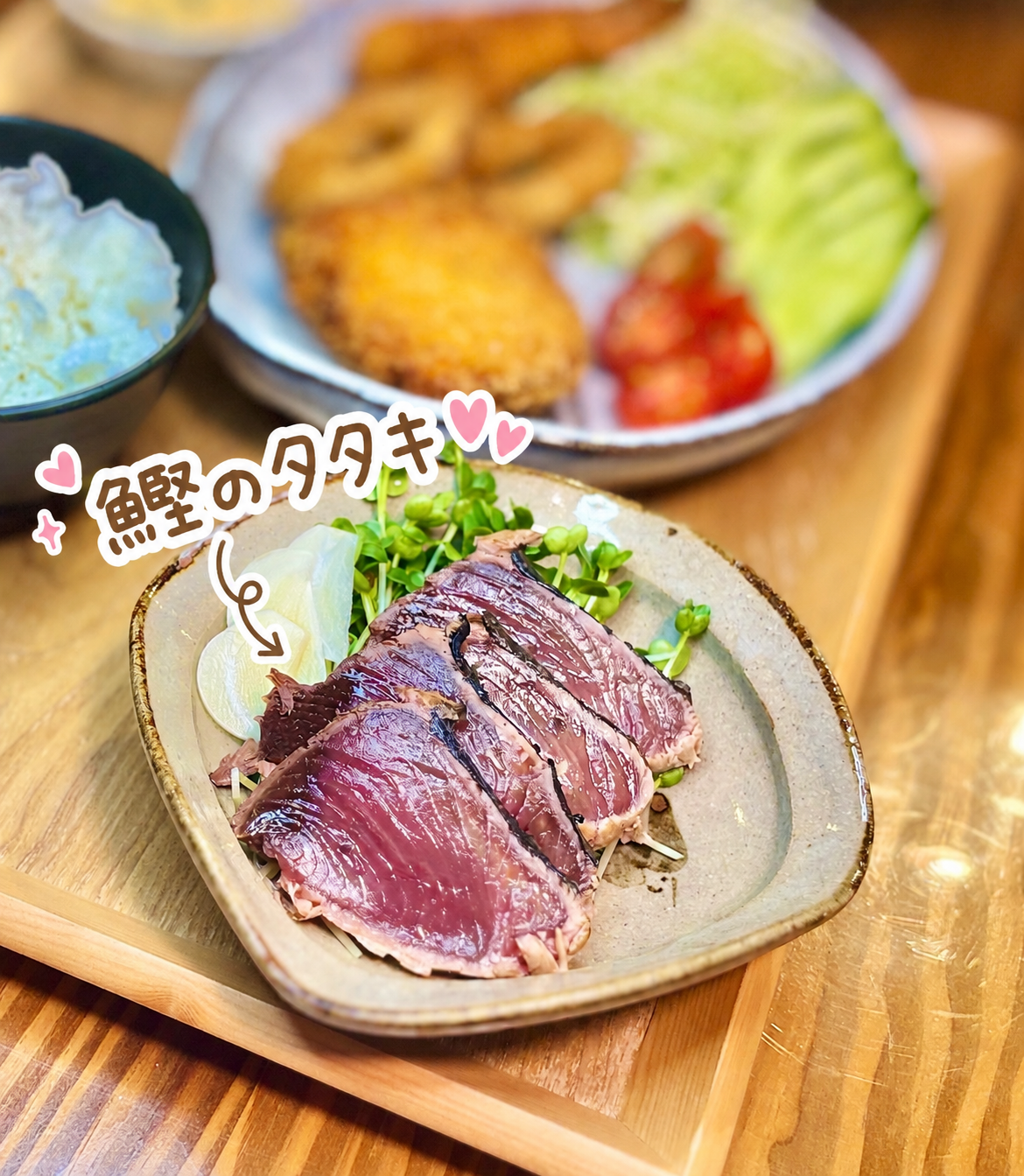 旦那キッチンの鰹のタタキのクローズアップ写真。表面を香ばしく炙ってロゼ色の断面が美しい鰹が、かいわれ大根とにんにくスライスを添えてベージュの角皿に盛り付けられている。奥にはブラックタイガーのエビフライ・イカリングフライ・クロカワカジキのフライと野菜サラダがぼんやり見える、揚げ物献立の副菜にもなる初鰹レシピ。