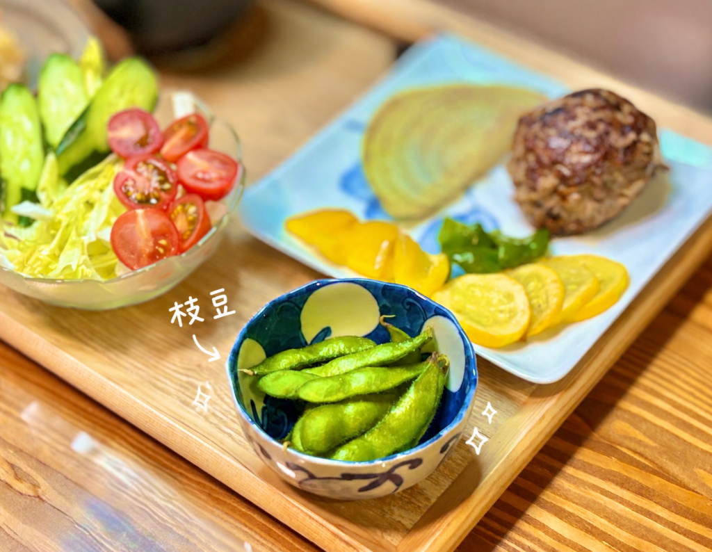 青と白の器に盛り付けた枝豆を、白い手書き風の文字と小さな星のイラストで可愛く紹介した写真。手前の枝豆はつやつやとした緑色がきれいで、奥にはハンバーグと焼き野菜、きゅうりとミニトマトのサラダが見えます。ハンバーグ献立に添える小さなおつまみや、食卓に彩りを加える副菜としてもぴったり。おうちごはん、晩ごはん献立、肉料理に合う簡単副菜を紹介する記事に使いやすい画像です。