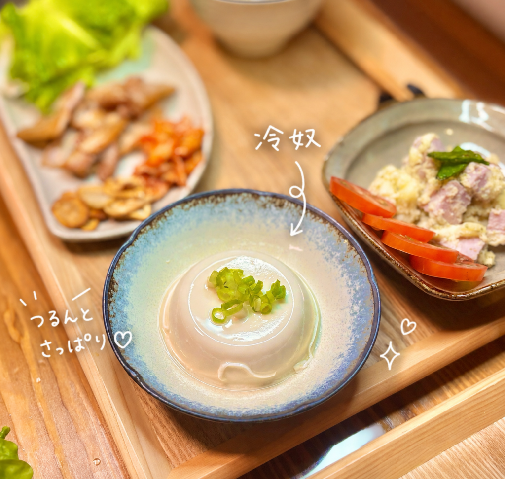 つるんとした冷奴に刻みねぎをのせ、国産豚バラ肉のサムギョプサルや明宝ハム紫蘇入り瑞峰のポテサラと一緒に木製トレーへ並べた副菜写真。豚バラ肉やキムチ、焼きにんにくのパンチがある献立の中で、冷奴が口の中をさっぱり整えてくれる一品として紹介されている。手書き風の文字入れで、冷奴の涼しげな雰囲気と家庭料理らしいほっとする副菜感を伝える画像。