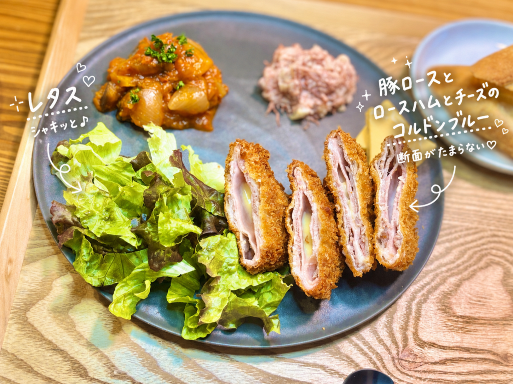 豚ロースとロースハムとチーズのコルドンブルーを食べやすくカットし、レタスと一緒に黒いプレートに盛り付けた洋風晩ごはんの一皿。サクッと香ばしい衣と、豚ロース・ロースハム・チーズが重なった断面が見えるコルドンブルーを、白い手書き文字で可愛く紹介した、おうちビストロ風のメイン料理写真。