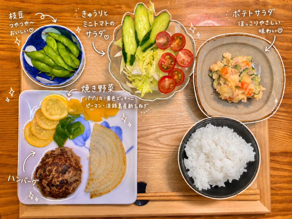 手作りハンバーグをメインに、焼き野菜、きゅうりとミニトマトのサラダ、ポテトサラダ、枝豆、白ごはんを木製トレーに並べたおうちごはん献立。焼き野菜にはパプリカ、黄色ズッキーニ、ピーマン、淡路島産新玉ねぎを添え、彩りよく仕上げています。写真には白い手書き風の文字入れで、ハンバーグ、焼き野菜、枝豆、サラダ、ポテトサラダを可愛く紹介。ハンバーグ定食の献立例や、肉料理に合う副菜を探している方にも参考になる、旦那キッチンの晩ごはん写真です。
