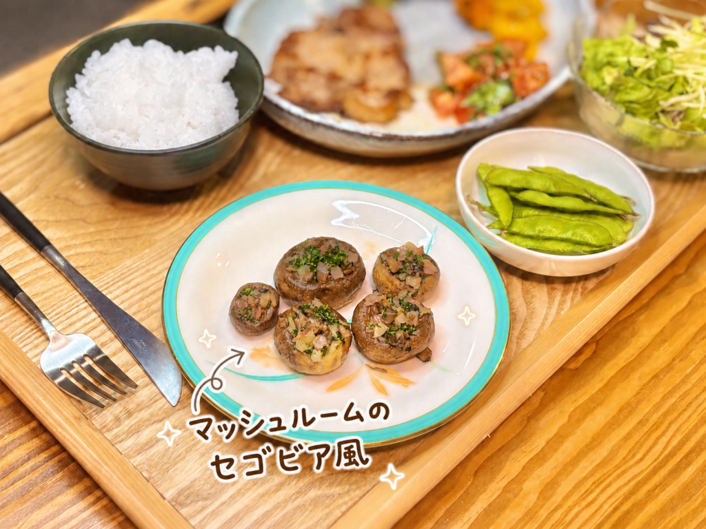 マッシュルームに刻んだ具材とハーブをのせて焼き上げた、マッシュルームのセゴビア風。木製トレーに並んだ晩ごはんの中で、前菜やおつまみにぴったりの一品を手書き風の文字入れで可愛く紹介した写真。