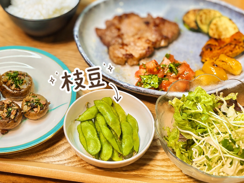 小鉢に盛り付けた鮮やかな緑色の枝豆を、手書き風の文字と矢印で可愛く紹介した一品写真。トンテキやマッシュルームのセゴビア風、野菜サラダと一緒に並ぶ、ビールに合う定番おつまみの枝豆。
