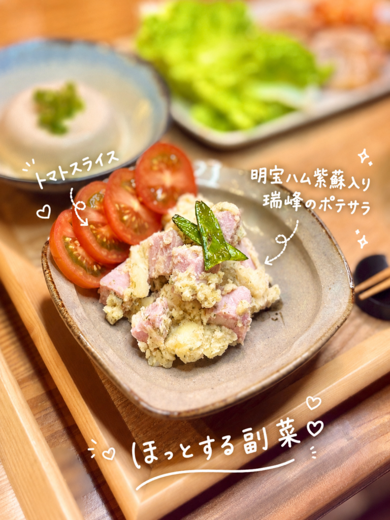 明宝ハム紫蘇入り瑞峰を使ったポテサラに、トマトスライスを添えた副菜の写真。ほくほくのじゃがいもに明宝ハムの旨みと紫蘇の香りを合わせたポテトサラダで、サムギョプサルのようなこってりした肉料理にも合うさっぱり副菜として盛り付けられている。手書き風の文字入れで、明宝ハム紫蘇入り瑞峰のポテサラとトマトスライスをわかりやすく紹介した旦那キッチンのおうちごはん画像。
