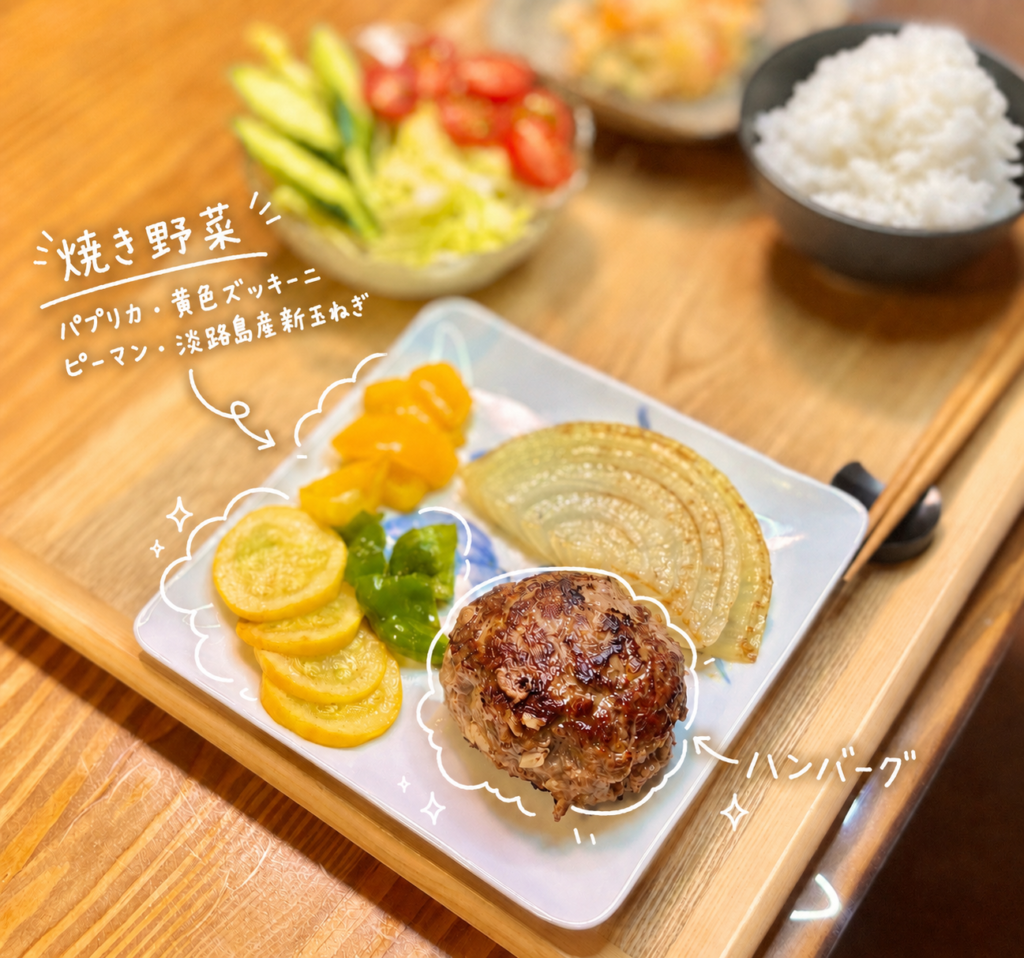 こんがり焼き色をつけた手作りハンバーグと、パプリカ、黄色ズッキーニ、ピーマン、淡路島産新玉ねぎの焼き野菜を盛り付けた一皿。手前のハンバーグは香ばしく焼き上がり、横には甘みのある新玉ねぎや鮮やかな焼き野菜を添えています。白い手書き風の文字入れで、焼き野菜の内容とハンバーグを可愛く紹介。おうちで作るハンバーグ献立、ハンバーグに合う付け合わせ、焼き野菜の盛り付けアイデアとして使いやすい写真です。