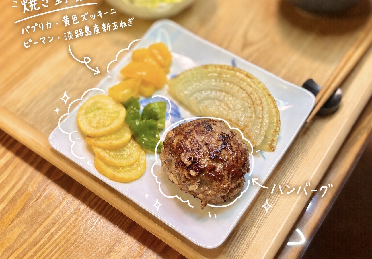 こんがり焼き色をつけた手作りハンバーグと、パプリカ、黄色ズッキーニ、ピーマン、淡路島産新玉ねぎの焼き野菜を盛り付けた一皿。手前のハンバーグは香ばしく焼き上がり、横には甘みのある新玉ねぎや鮮やかな焼き野菜を添えています。白い手書き風の文字入れで、焼き野菜の内容とハンバーグを可愛く紹介。おうちで作るハンバーグ献立、ハンバーグに合う付け合わせ、焼き野菜の盛り付けアイデアとして使いやすい写真です。