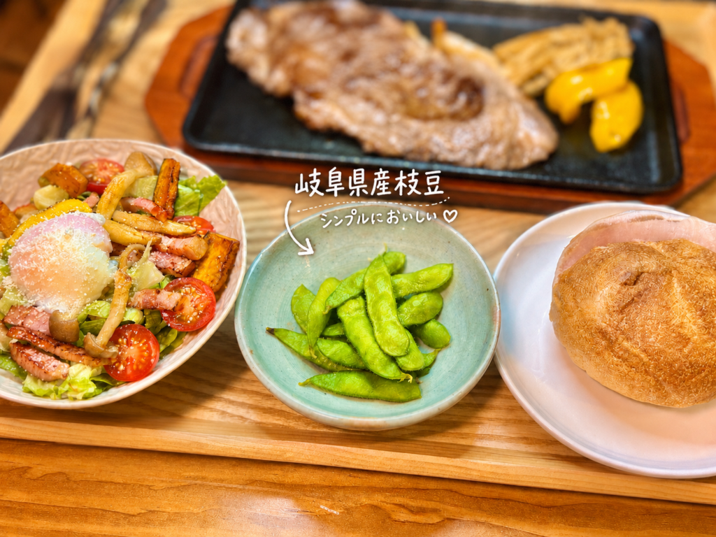 岐阜県産枝豆を小鉢に盛り付けた、おうちごはんの副菜写真。木製トレーの上に、具沢山シーザーサラダ、ダンマルシェのカイザーパン、奥にはロースステーキの鉄板プレートを並べ、中央に鮮やかな緑色の枝豆を配置した献立の一枚。ロースステーキや赤ワインに合わせる食卓で、シンプルながら箸休めにもなる枝豆を添えた旦那キッチンのごちそうディナー。