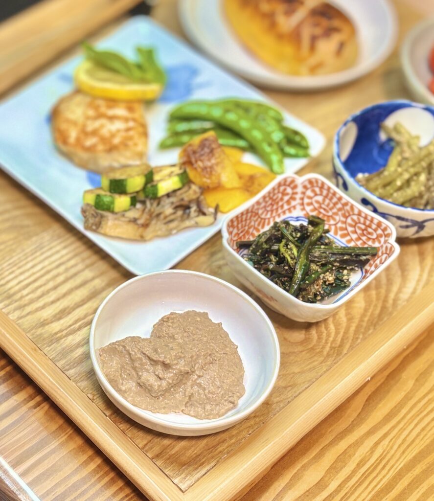 鳥レバーのパテ、わらびのナムル、ふきのナムル、奥に真鯛のムニエル レモンバターソースと焼き野菜、コープの塩バターチーズパンを並べたおうちビストロ風献立の食卓写真。