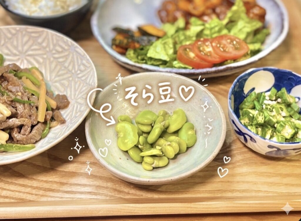 旬のそら豆を塩茹でにした副菜のアップ写真。鮮やかな緑色のそら豆が素朴なグレーの小鉢に盛り付けられている。周囲には青椒肉絲・おくらポン酢・酢豚・トマトサラダが並ぶ旦那キッチンの晩ごはん食卓。ほくほく食感が楽しめる旬野菜の簡単副菜レシピ。