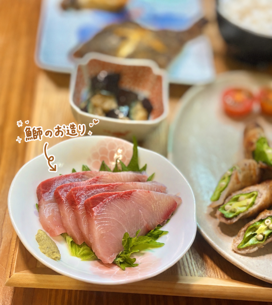 鰤のお造りを白い花柄のお皿に盛り付けた写真。大葉の上に並べた脂ののった鰤の切り身とわさびを添え、手書き風の文字で料理名を入れた、旦那キッチンの刺身料理画像。