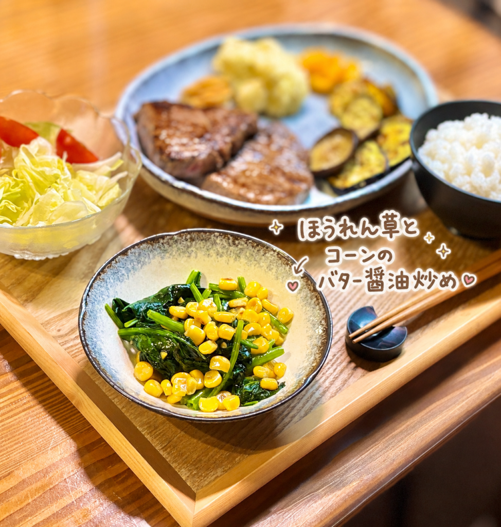 ほうれん草とコーンのバター醤油炒めを小鉢に盛り付けた写真。奥には和牛ランプステーキや野菜サラダ、白ごはんが見え、手書き風の文字で副菜名を紹介した、彩りのよい付け合わせ画像。