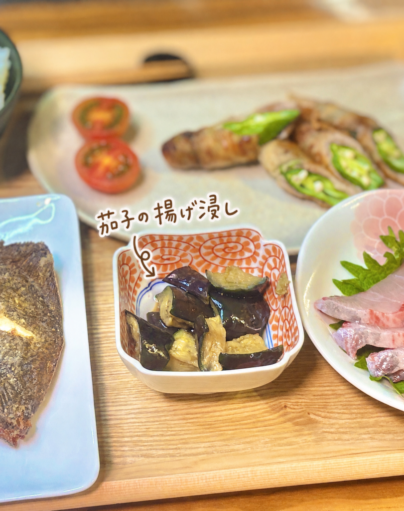 茄子の揚げ浸しを小鉢に盛り付けた写真。手書き風の文字と矢印で料理名を入れ、奥にはおくらの豚バラ巻きや鰤のお造り、メイタガレイの唐揚げが見える、旦那キッチンの和食副菜画像。